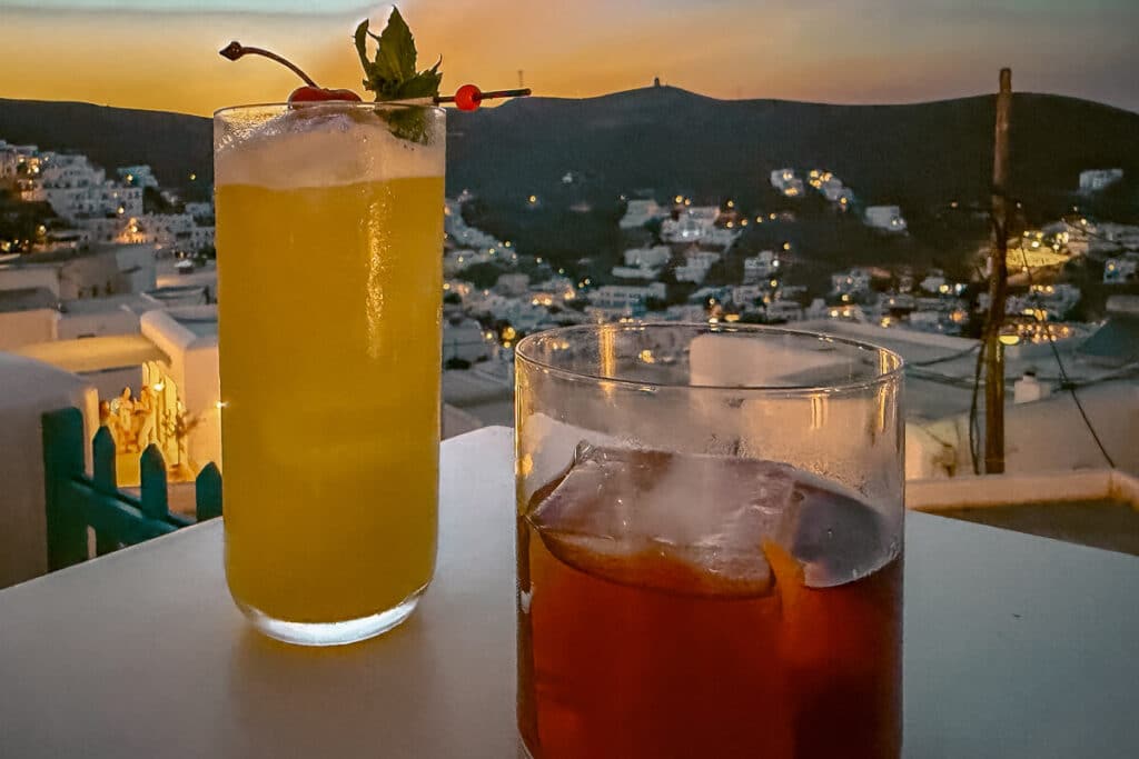 Two cocktails on a rooftop table at sunset in Astypalaia, with whitewashed Chora buildings and glowing evening lights in the background.