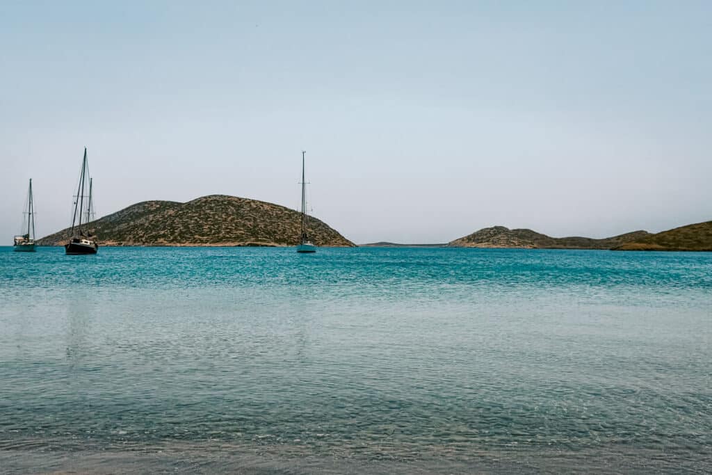 Clear turquoise water at Paralia Schinonta in Astypalaia, with sailboats floating offshore and dry rocky islets in the distance.