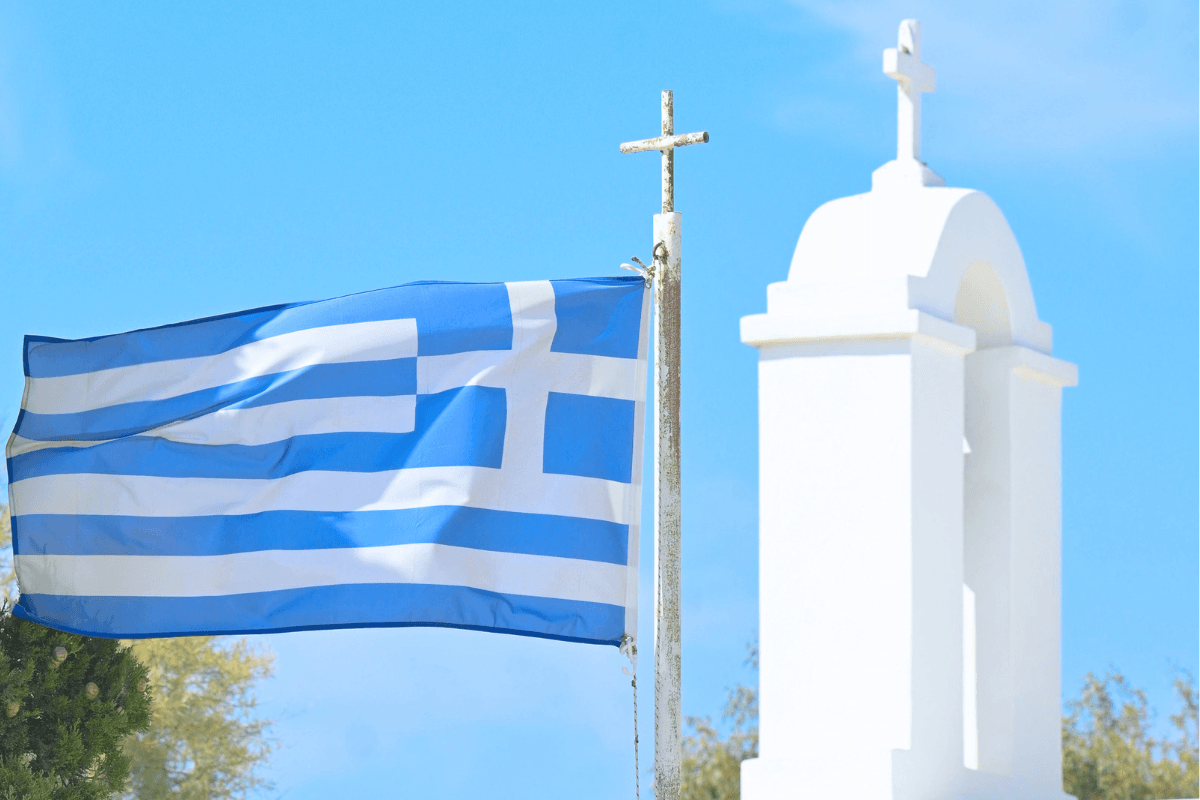 Greek flag waving beside a traditional white church against a blue sky, representing basic Greek words for travelers visiting Greece.