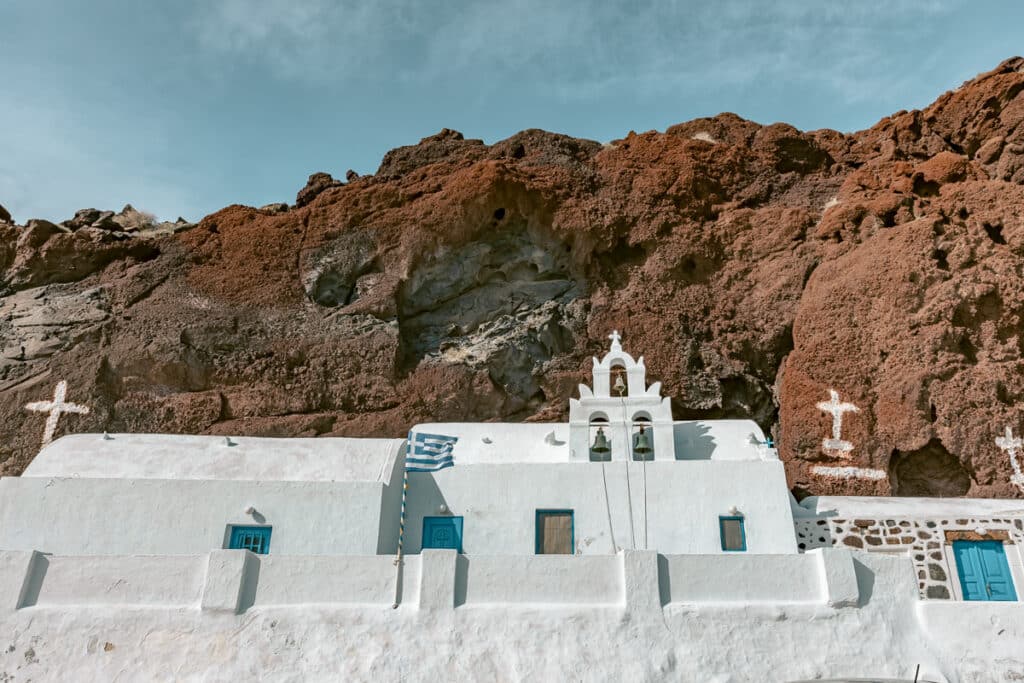 White church with blue doors built below the red volcanic cliffs at Red Beach in Santorini, a scenic stop to add to a Greece itinerary.