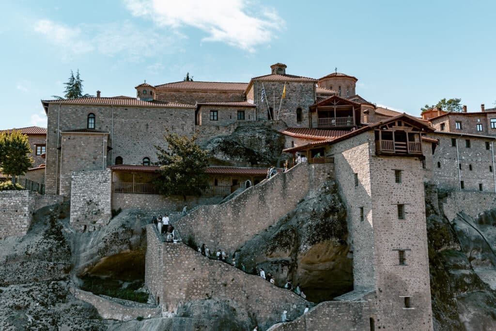 Stone monastery built into the rock formations of Meteora, Greece, with visitors walking along the staircase entrance.
