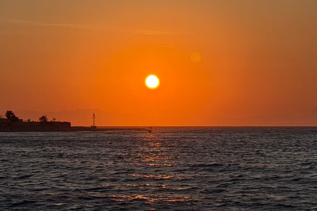 Orange sunset over the sea in Rhodes with the coastline in the distance, a scenic evening view to include in an Athens, Rhodes, and Symi 1 week in Greece itinerary.