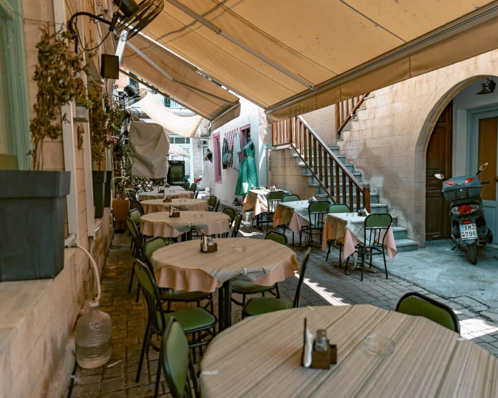 Narrow alleyway in Aegina with round café tables under a yellow awning, decorated with dried herbs, plants, and colorful local charm.