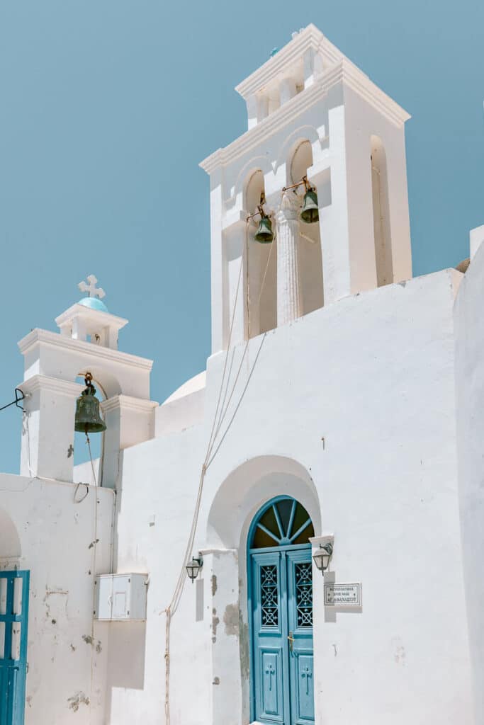 Bright white Greek island church with a blue entrance door and two bell towers under a clear blue sky.