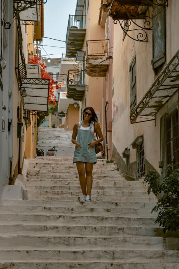 standing on a long staircase in Nafplio surrounded by pastel-colored buildings, red flowers, and charming old balconies.