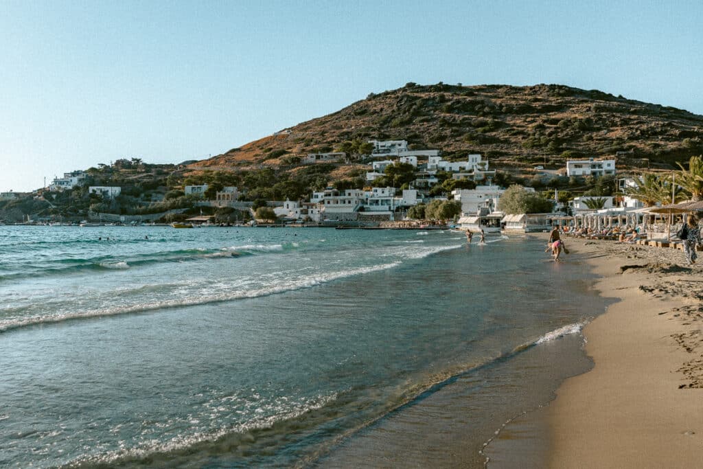 A sandy beach in Syros with gentle waves lapping at the shore, sunbeds lined along the right side, and whitewashed houses climbing the hillside in the background.