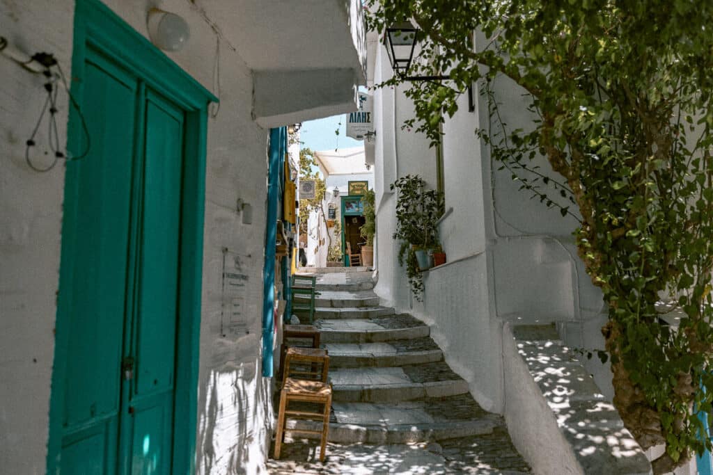 A narrow alleyway in Syros filled with potted plants and colorful doors, shaded by leafy trees and framed by bright white Cycladic walls. A tucked-away spot perfect for wandering during a 1 week in Greece itinerary.