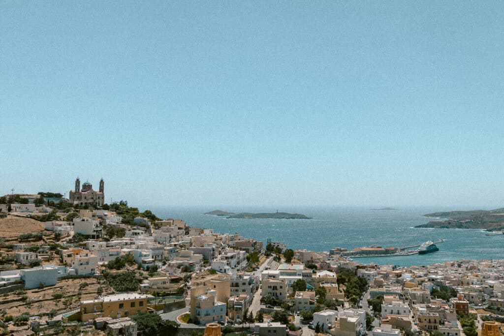 A panoramic view from of Syros, with a harbor, distant islands on the horizon, and a prominent church perched on a hill. This wide shot shows how coastal towns blend natural beauty with classic Greek architecture.