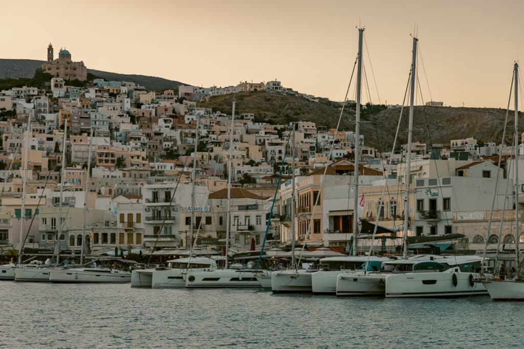 A cluster of sailboats docked in a harbor at sunset, with pastel homes stacked on the hillside behind them. This scenic harbor view in Syros brings a calm close to a day exploring Greece on a 7 day itinerary.