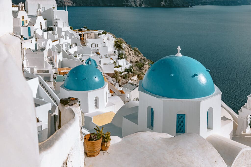 Whitewashed buildings and two bright blue church domes overlooking the sea in Oia, Santorini, with cliffs and deep blue water stretching into the distance.