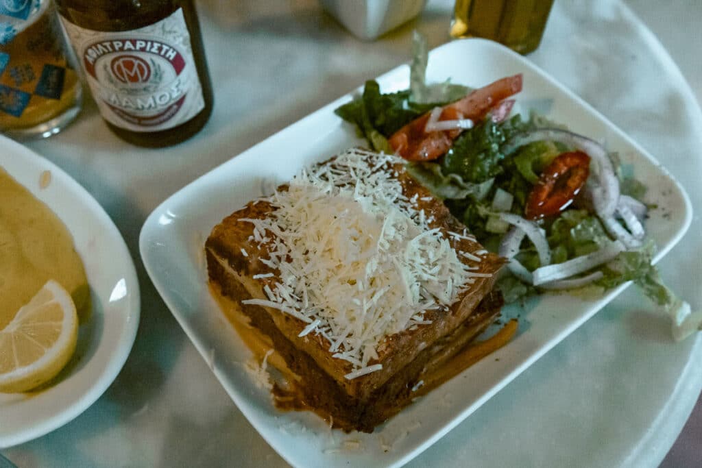 Classic Greek patstitio with grated cheese on top, served with a simple salad and a bottle of Mamos beer in the background. Indoor taverna setting with cozy lighting.