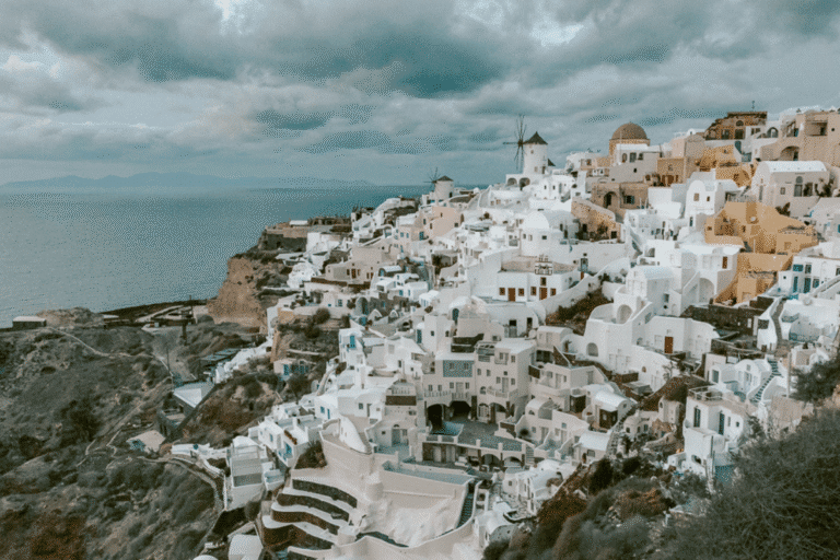Whitewashed buildings and traditional windmills in Oia overlooking the Aegean Sea under dramatic clouds, featured in a collection of Santorini captions.