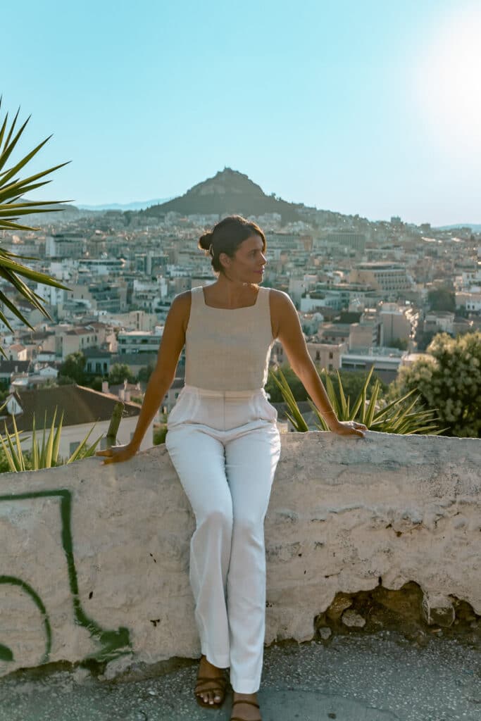Me sitting on a low stone wall overlooking the Athens skyline, with Mount Lycabettus in the distance. The golden hour light and her calm expression capture the magic of discovering the city solo.