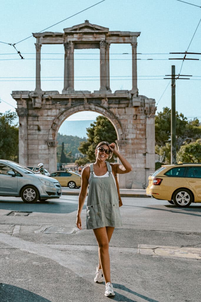 Me grinning playfully in front of Hadrian’s Arch in Athens, surrounded by passing traffic and yellow taxis. She’s adjusting her sunglasses mid-walk, embracing the energetic chaos of city life.