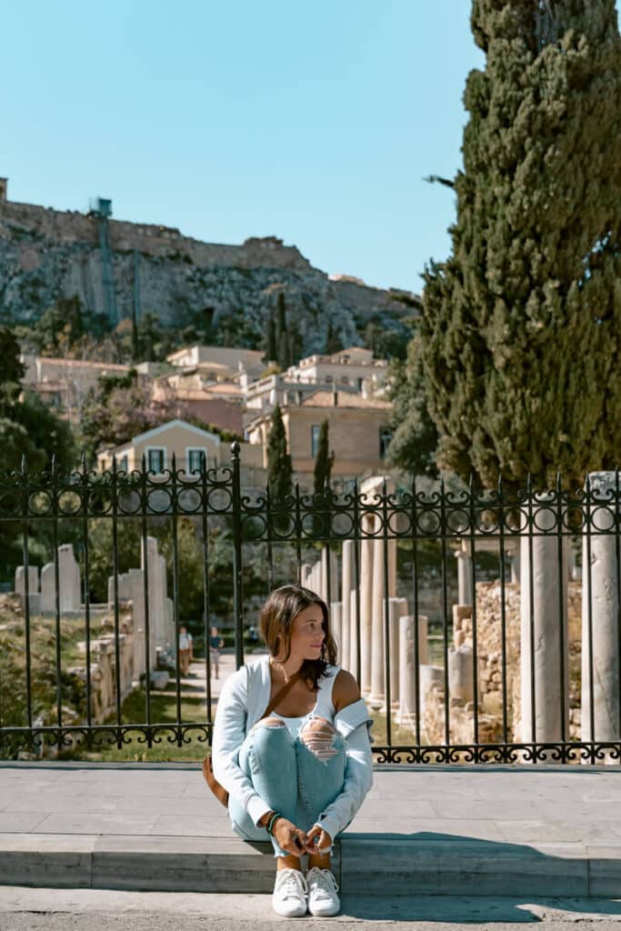 Me sitting cross-legged on a stone step in front of a wrought-iron fence with ancient columns behind her in Athens. She's looking to the side with a relaxed smile, dressed casually in ripped jeans and a light jacket. The Acropolis rises in the background, adding a historical feel to this peaceful moment of solo travel.