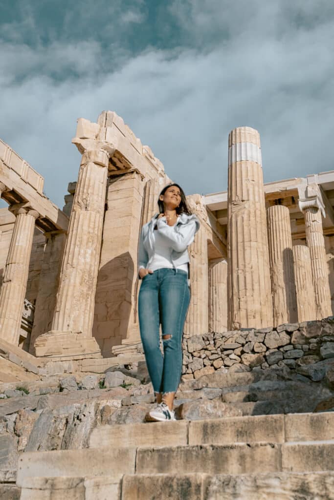 Me standing at the Acropolis in Athens, standing confidently in front of ancient stone columns with one hand in her pocket and the other brushing her hair. The dramatic sky adds contrast to the scene, reflecting the sense of awe that often comes with solo travel to Athens.