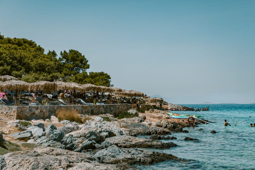 Aerial view of turquoise water and rocky coastline lined with straw umbrellas, one of the top relaxing things to do in Agistri, Greece.