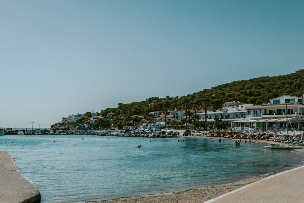 A calm bay in Skala with turquoise water, people swimming, and sunbeds lined up along the shore, backed by charming white buildings and green hills.