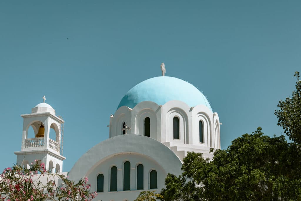 A stunning Greek Orthodox church with a bright blue dome and whitewashed bell tower under a clear blue sky, framed by lush green trees and pink flowers.