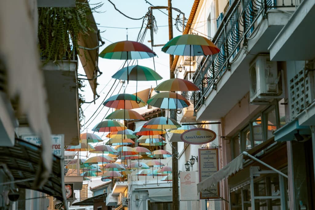 Colorful umbrellas suspended above a narrow shopping street in Aegina, Greece, adding a playful and artistic touch to a pedestrian walkway lined with local shops.