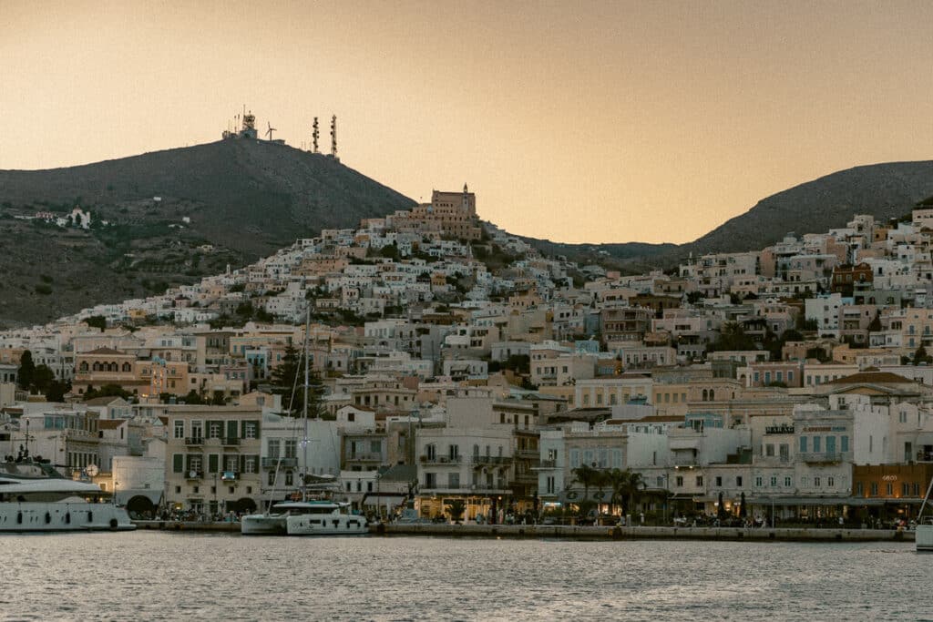 Harbor town in Syros, Greece at sunset with layered stone buildings rising up a hill and boats docked along the waterfront, creating a warm evening atmosphere for a 5 day Greece itinerary.