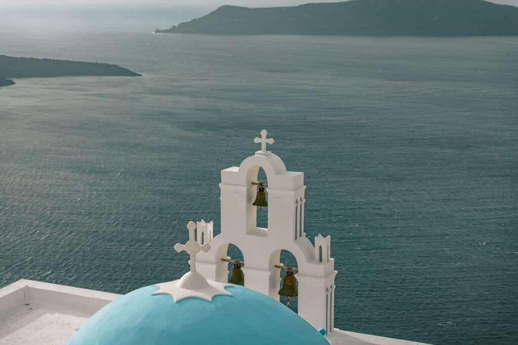 White bell tower and blue dome church overlooking the Aegean Sea in Santorini, with volcanic cliffs and open water stretching into the distance.