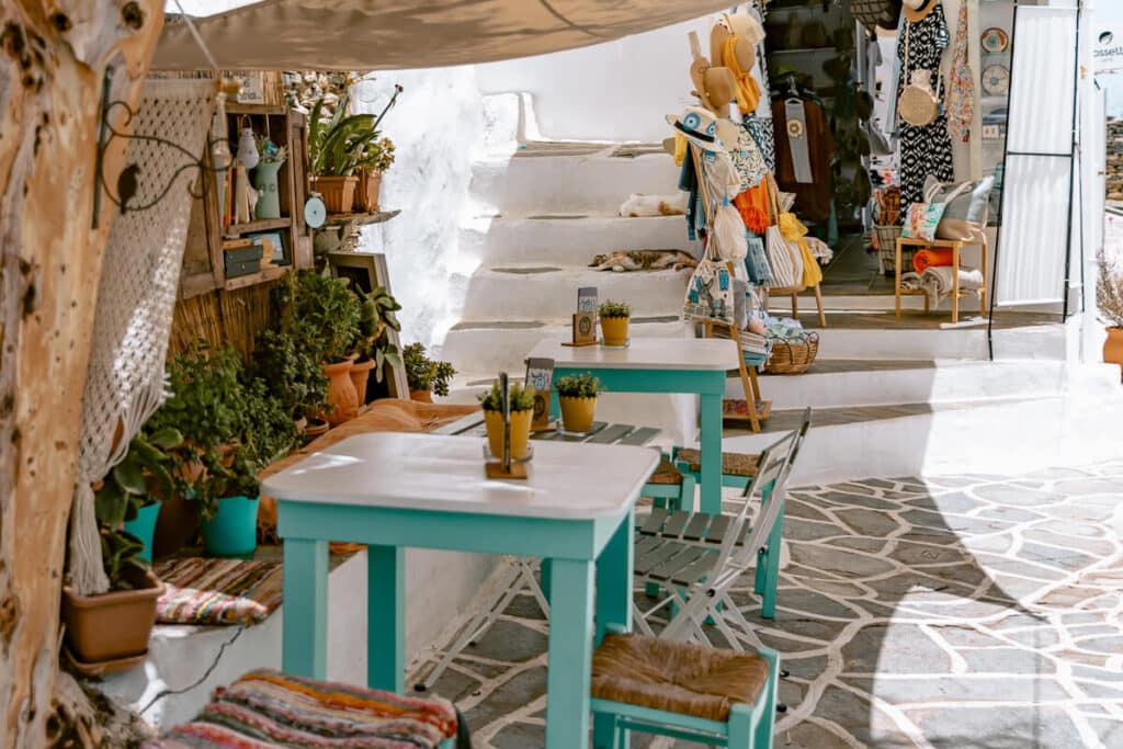 Small boutique shop on Sifnos with whitewashed walls, handmade textiles, pottery, and turquoise tables set along a stone walkway, showing everyday island life and local craftsmanship.