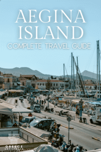 Busy Aegina Island harbor with boats, scooters, and shops, set against a mountainous backdrop, representing the lively port area. Text overlay reads: “Aegina Island – Complete Travel Guide.”