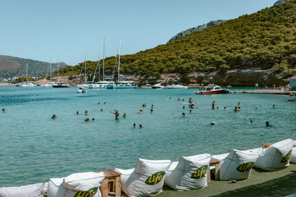 Moni Island with clear turquoise water, swimmers in the sea, and sailboats anchored nearby.