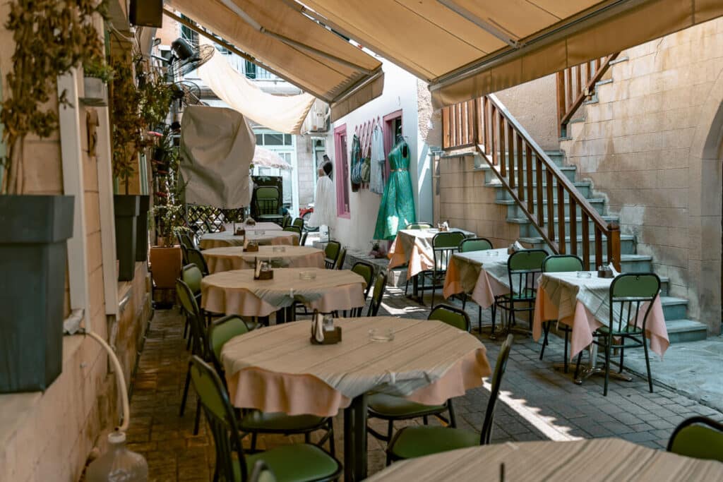 Outdoor taverna seating in a shaded alley in Aegina with small tables and chairs, showing a relaxed dining scene.