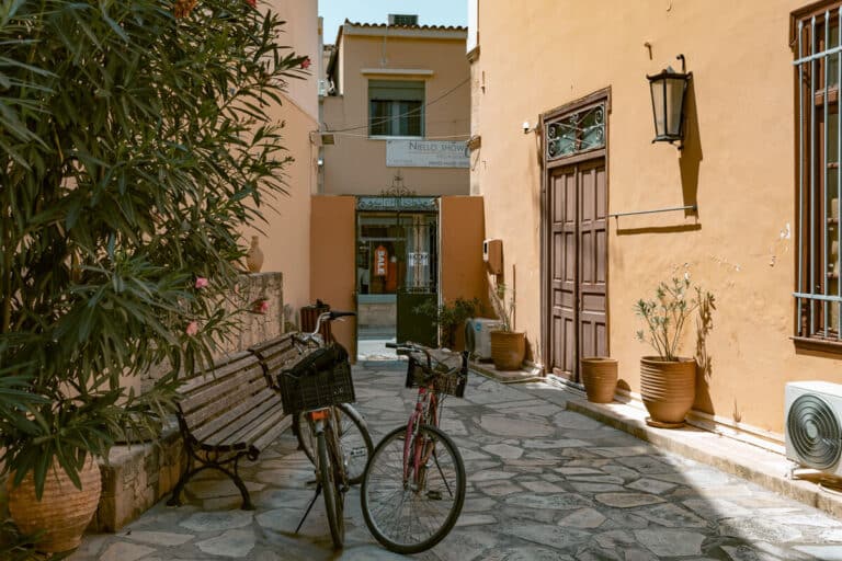 Bicycles parked along a quiet stone paved street with warm colored buildings and potted plants during a day trip to Aegina.