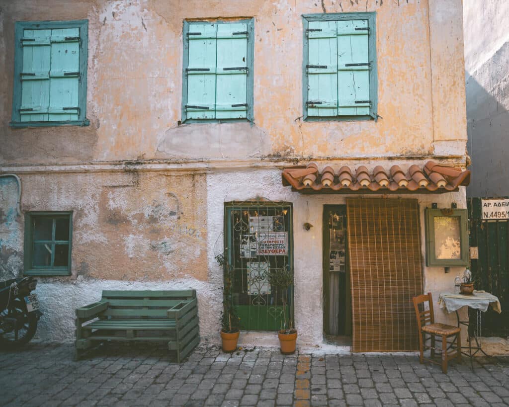 Weathered traditional building in Aegina with faded peach walls, turquoise wooden shutters, a tiled awning, and a small table and chair outside.
