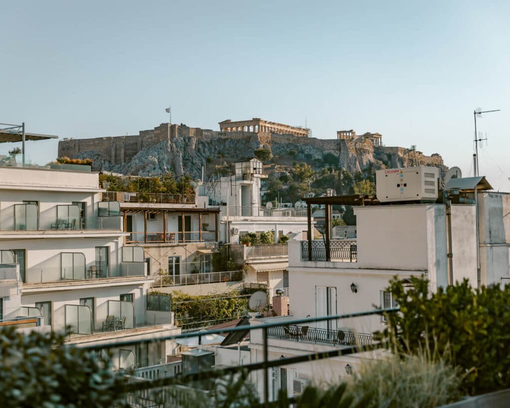 Rooftop perspective of modern buildings with the Acropolis and Parthenon perched above, seen in late-day light.