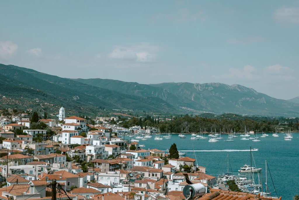 Harbor town with terracotta rooftops and sailboats anchored in calm blue water against a mountain backdrop in Poros.