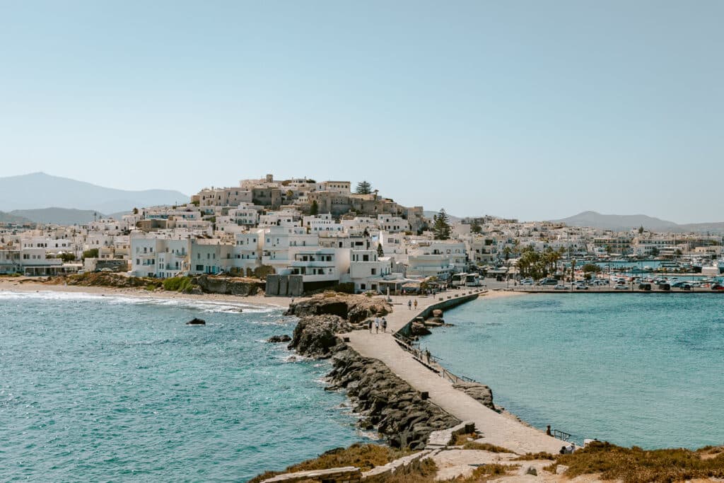 Panoramic view of Chora, Naxos, with whitewashed hillside buildings, the harbor, turquoise water, and a stone walkway curving along the coast.