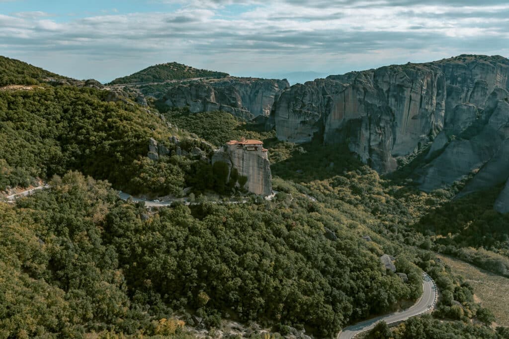 Panoramic view of Meteora with towering rock formations, winding roads, forested hills, and a monastery perched high on the cliffs.