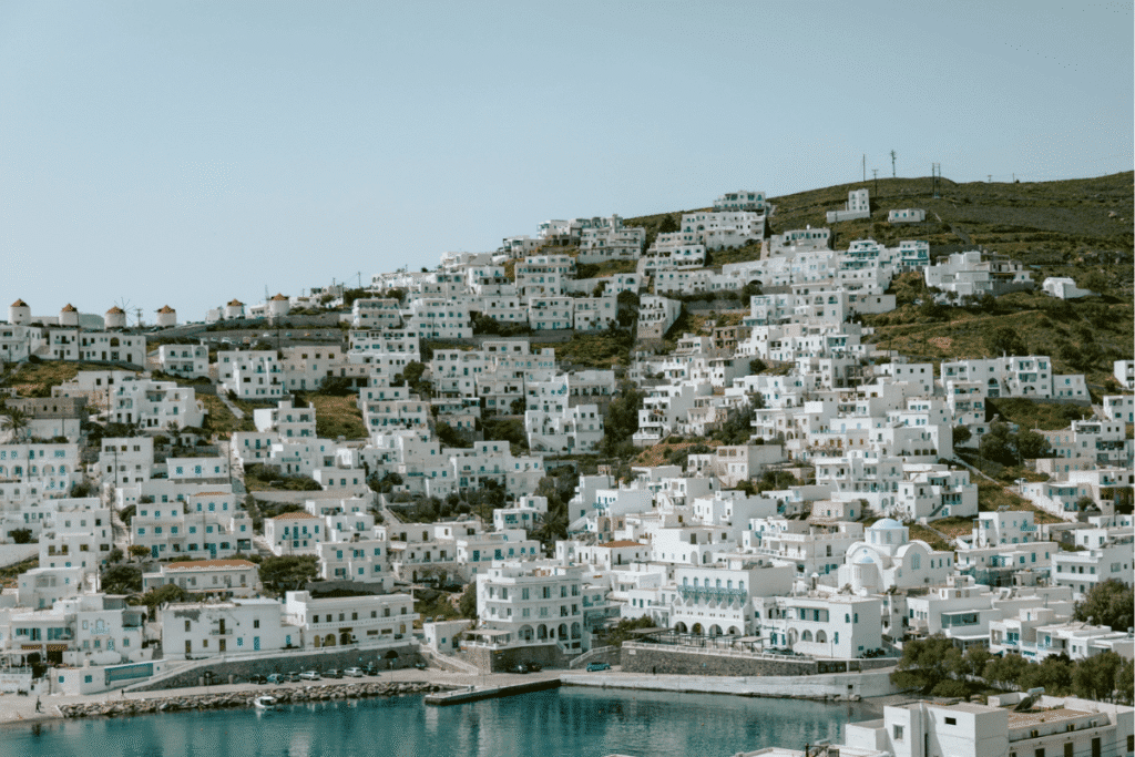 Whitewashed hillside village with blue shutters, traditional windmills, and a waterfront overlooking the Aegean Sea, showcasing the scenery found on the best Greek islands to visit for the first time.