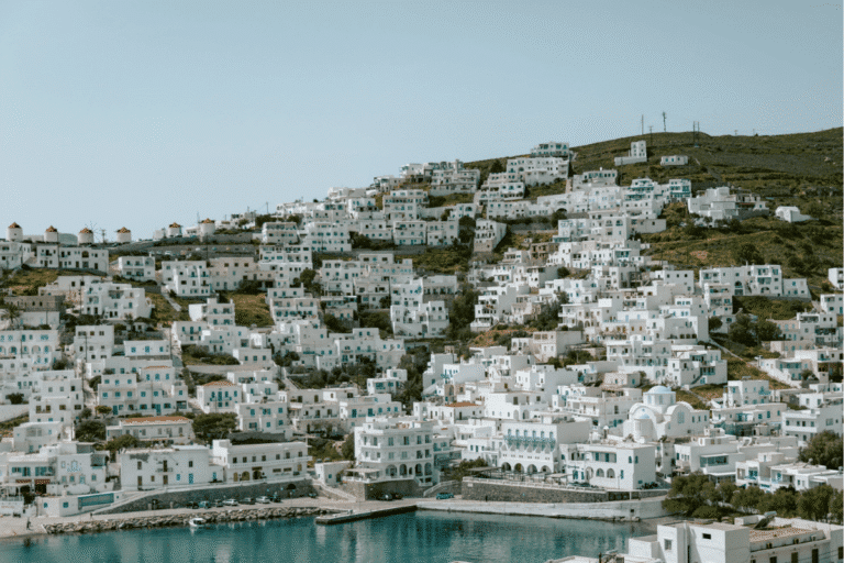 Whitewashed hillside village with blue shutters, traditional windmills, and a waterfront overlooking the Aegean Sea, showcasing the scenery found on the best Greek islands to visit for the first time.