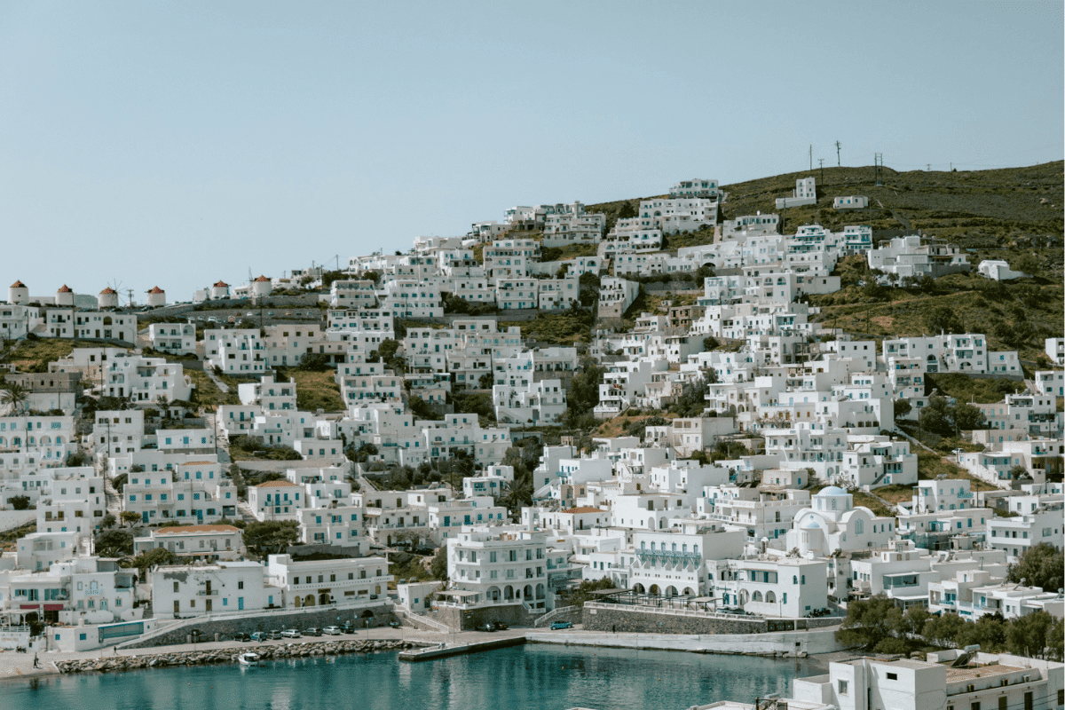 Whitewashed hillside village with blue shutters, traditional windmills, and a waterfront overlooking the Aegean Sea, showcasing the scenery found on the best Greek islands to visit for the first time.
