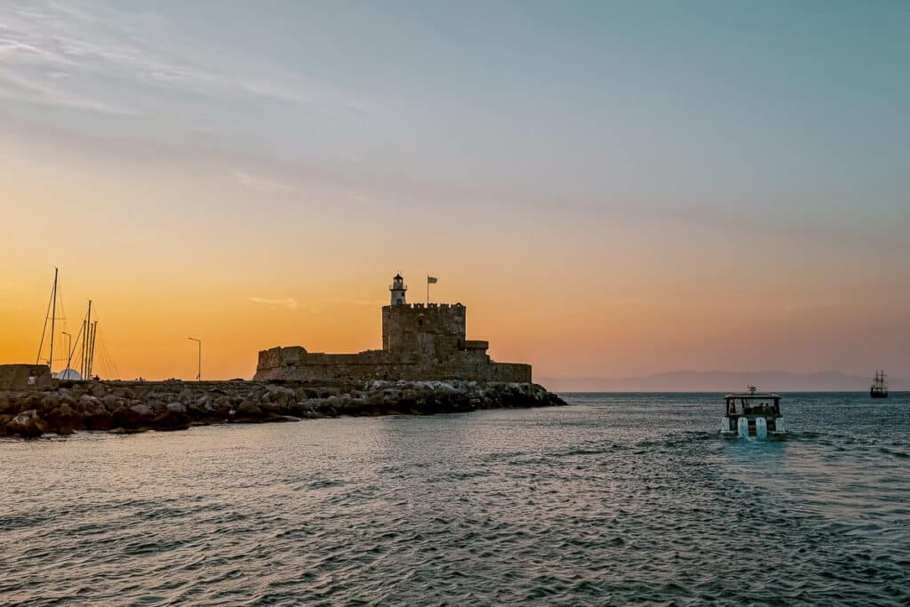 Sunset over the harbor in Rhodes, Greece with the medieval St. Nicholas Fortress by the water, one of the best Greek islands to visit for the first time.