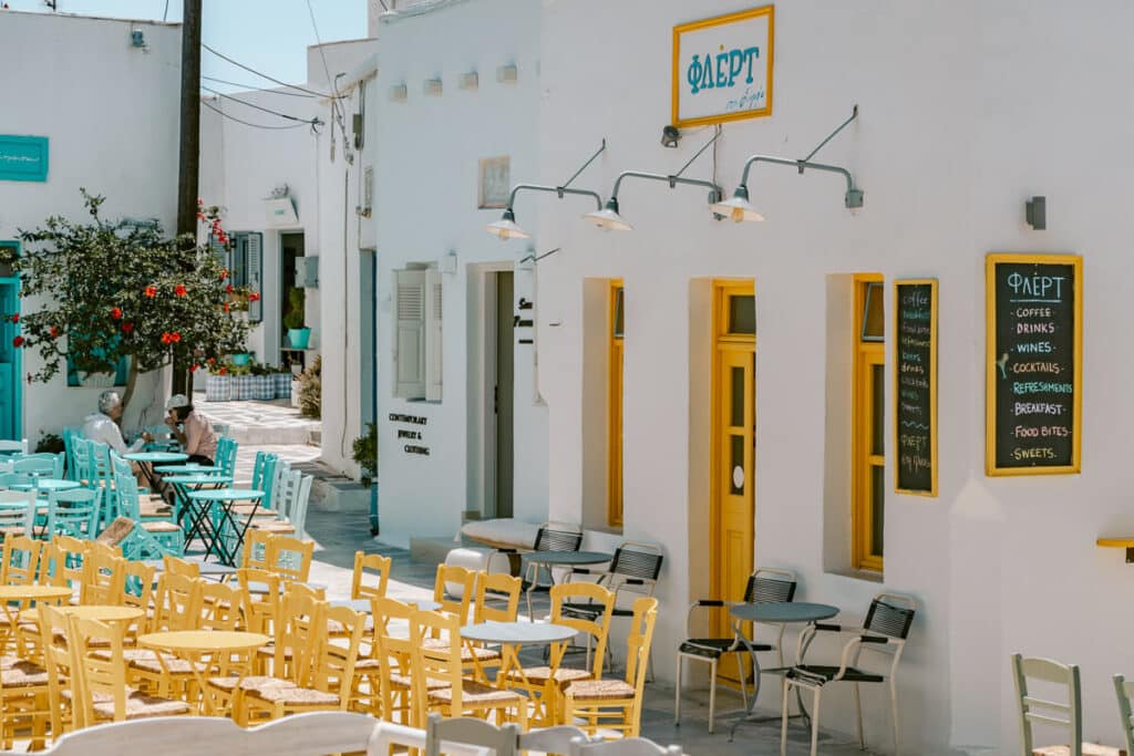 Outdoor café in a whitewashed Greek village with yellow chairs and tables lined along a sunny lane.