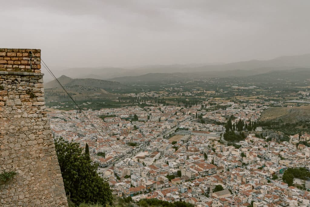 Wide view over Nafplio from above, showing red tiled rooftops, rolling hills, and a hazy sky