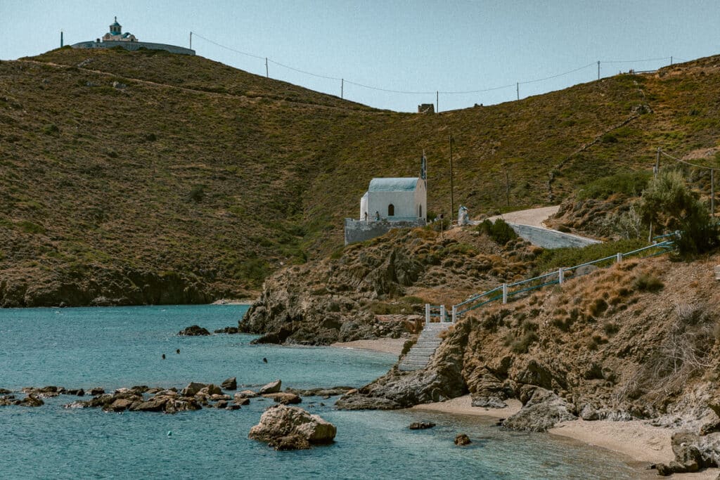 Small beach cove in Greece with clear turquoise water, rocky shoreline, and tiny white chapels on the hills above.