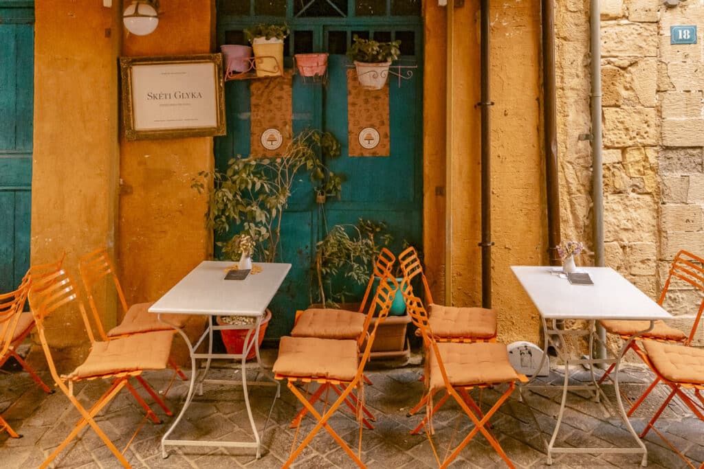 Small outdoor café with orange chairs and white tables set against warm stone walls in a narrow old town street in Greece.