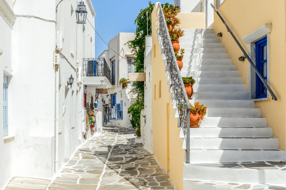 Sunny Greek island lane with whitewashed walls, a pale yellow staircase, and blue accents, capturing the best time to visit Greece for warm weather and quiet village charm.
