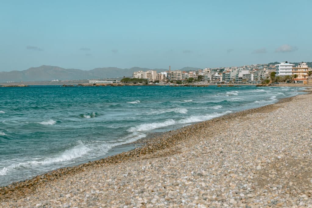 Pebble beach with turquoise water and seaside buildings in Chania, showing one of the coastal views near the city