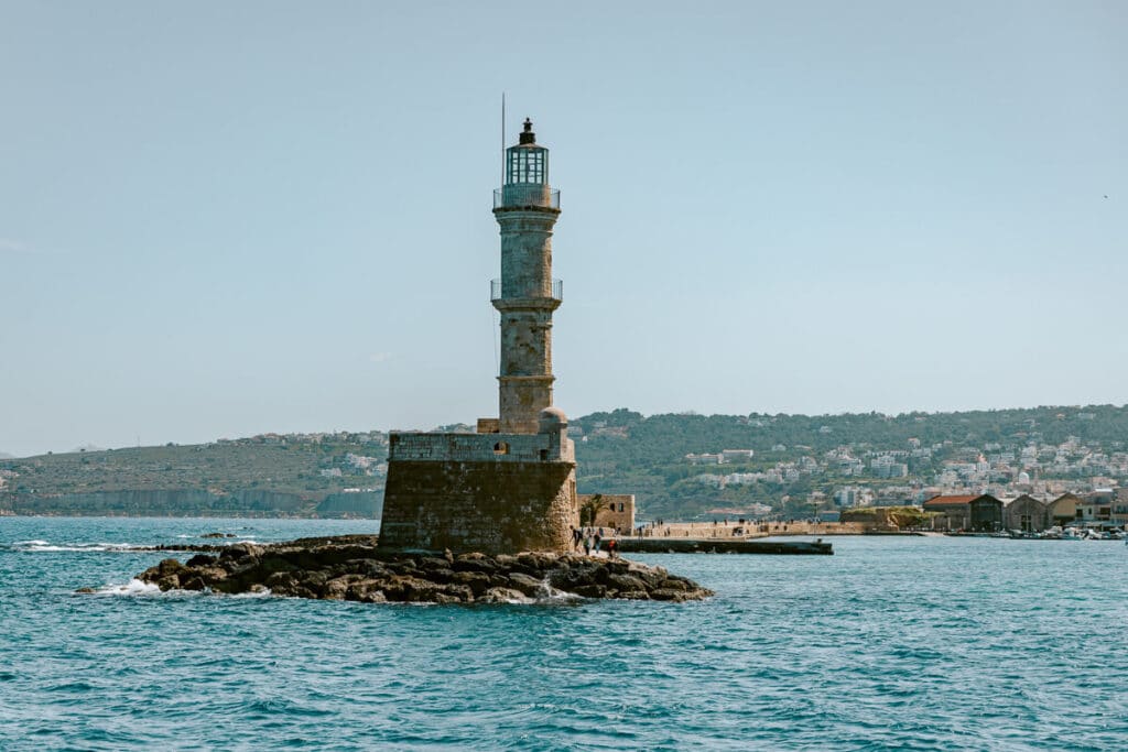 Closer view of the Chania lighthouse surrounded by blue water, with the harbor and hillside in the background