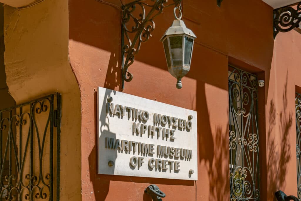 Sign for the Maritime Museum of Crete on a terracotta wall in Chania, with wrought iron window details and a hanging lantern