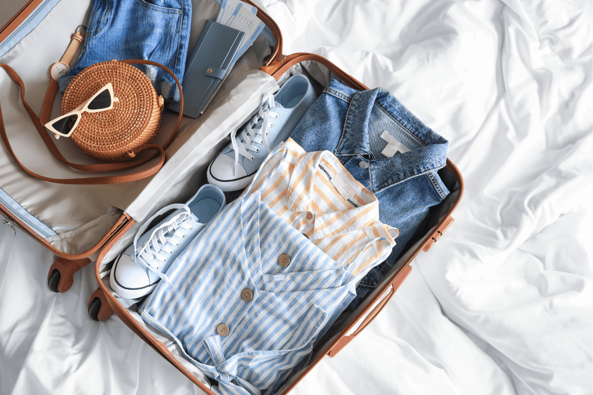 Open suitcase packed with sneakers, denim, striped clothing, sunglasses, travel documents, and a woven bag for an Athens packing list.