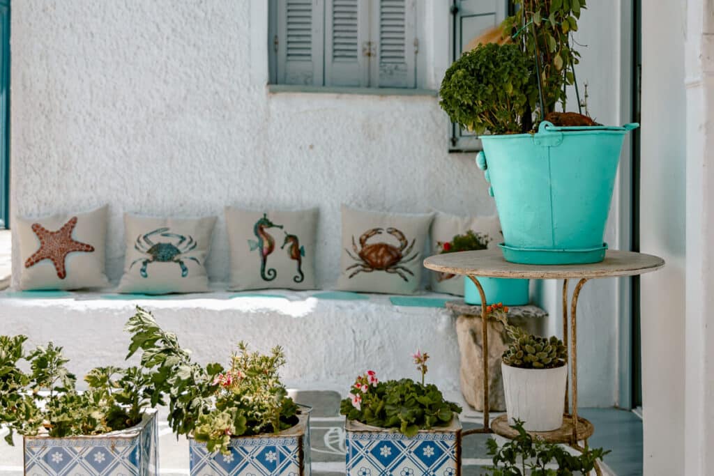 White Greek building with blue shutters, sea themed pillows, and potted plants, showing the simple island charm travelers can enjoy while planning Greece on a budget.